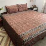 Rust red floral Kalamkari double bedsheet with indigo and mustard border on a bed with two pillow covers